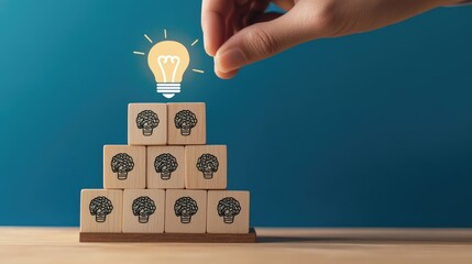 A hand places a wooden block with a lightbulb icon on top of a pyramid of blocks depicting heads with brain icons on a wooden surface against a blue background.