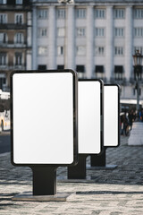 Modern urban street advertising displays or billboards with blank white screens positioned in a row on a tiled pavement. City buildings in the background, no visible advertisements, ideal for mockups