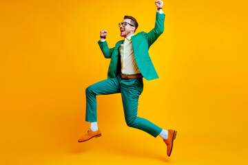 Young man celebrating success. Happy funny joyful excited guy in stylish green party suit and cool glasses raising fist up and dancing isolated on bright yellow background. Full length shot, side view