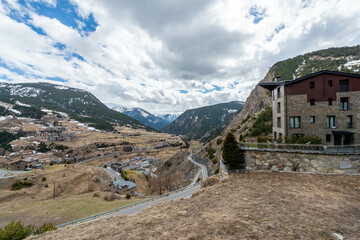 A winding road leads through a Pyrenean valley towards a quaint town nestled below snow-capped mountains, with a traditional stone building overlooking the scene under a partly cloudy sky.