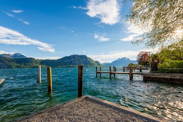 Peaceful Lake Lucerne waterfront with wooden pier, mountains and blue sky on sunny day.