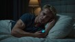 © theartofphoto - Attractive young blond man lying in bed at night holding a device, pondering under soft lamplight.