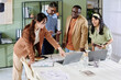 © pressmaster - Group of diverse colleagues engaging in collaborative discussion around office table, surrounded by laptops, documents, and stationary, planners examining financial reports together