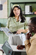 © pressmaster - Woman holding tablet presenting marketing data to colleague in office environment. Engaged in discussion while standing by desk with laptop, papers, and various office supplies
