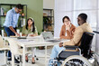 © pressmaster - Group of colleagues working together in office, including person in wheelchair, engaging in discussion around laptops on table during meeting