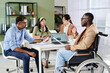 © pressmaster - Group sitting around table in office discussing strategies while one man in wheelchair presenting data using laptop engaging team members in conversation