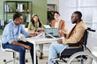 © pressmaster - Group of business professionals sitting around table, engaging in important meeting. People reviewing documents, one person in wheelchair, modern office environment