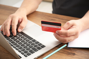 Man with credit card using laptop at wooden table indoors, closeup