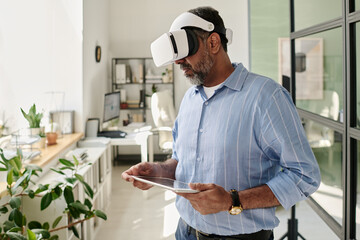 Bearded man using virtual reality headset while holding tablet in airy office with plants and large windows, deeply engaged with interactive virtual content