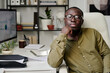 © pressmaster - Portrait of an African American man sitting at modern office setup, wearing glasses and green shirt, with thoughtful expression, surrounded by office supplies and indoor plants