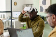 © pressmaster - Man engaging with virtual reality headset, sitting at desk with laptop in modern office featuring coworkers and tech setup in background