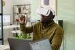 © pressmaster - Man sitting at desk wearing virtual reality headset, interacting with virtual elements. Modern office setting visible with shelves and plants creating a realistic backdrop