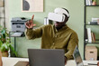 © pressmaster - Person engaging with virtual reality in an office environment with bookshelves and printer in background while seated at desk.