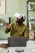 © pressmaster - African American man wearing virtual reality headset and sitting at desk with laptop, raising hand as if interacting with virtual environment