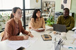 © pressmaster - Asian woman in glasses leading presentation with notebook while colleagues listen attentively in modern office with large windows and various office supplies on desk