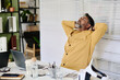 © pressmaster - Businessman relaxing at work with arms stretched behind head. Modern office setting involving various gadgets and devices visible on desk without usage of natural light