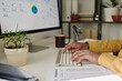 © pressmaster - Person typing on keyboard while analyzing data on computer screen and reviewing printed report. Desktop decorated with potted plants and a brown coffee mug