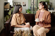 © pressmaster - Two Asian women sitting on a couch while conversing and enjoying coffee in well-designed living room setting with plants and modern decor