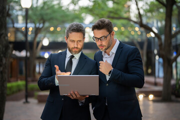 Two businessman on the street, deeply engrossed, eyes fixed on their laptop screen. Two handsome young businessmen in classic suits using laptop. Business men team using laptop outdoor. Business man