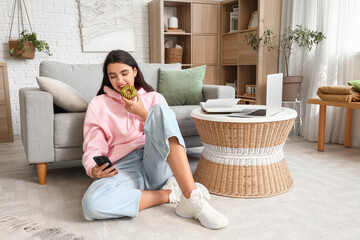 Canvas Print - Beautiful young woman in stylish hoodie with donut and mobile phone sitting at home