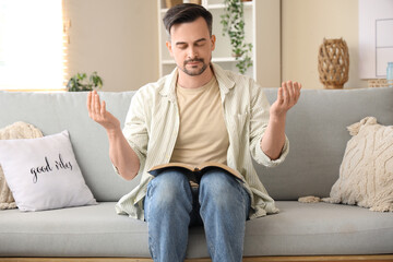 Canvas Print - Religious young man with Holy Bible praying on sofa at home