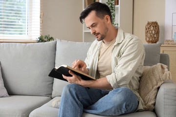 Wall Mural - Religious young man reading Holy Bible on sofa at home