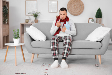 Canvas Print - Ill young man with hot water bottle and thermometer measuring temperature on sofa at home