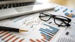 © improvee design - Close-up of a desk with a laptop, glasses, pen and financial data charts, suggesting business planning and analysis.