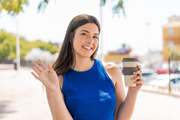 Young pretty Brazilian woman holding a take away coffee at outdoors saluting with hand with happy expression