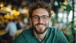 © JoxyAimages - Bearded man in glasses smiles brightly while seated in a cozy cafe environment, depicting warmth, friendliness, and a relaxed atmosphere in modern urban life.