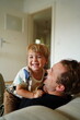 © Татевик Багдасарян - A smiling young boy sitting on his father's lap, both sharing a joyful and playful moment together at home.