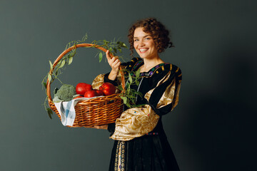 Wall Mural - Portrait of a young adult woman dressed in a medieval dress holding a basket with vegetables and fruits, apples and brussels sprouts. Harvest concept.