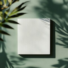 A minimalist square notebook sits on a green surface, with shadows from surrounding plants casting intricate patterns that enhance the tranquil atmosphere.