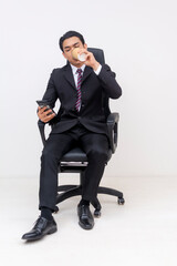 Young Asian businessman in suit using phone and drinking coffee. Whole body photo.