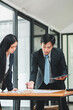 © Satori Studio - Two business professionals in formal attire discussing documents at a table in a modern office environment.