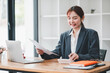 © Satori Studio - A young professional woman in a business suit working at her desk with a laptop and documents in a modern office environment.