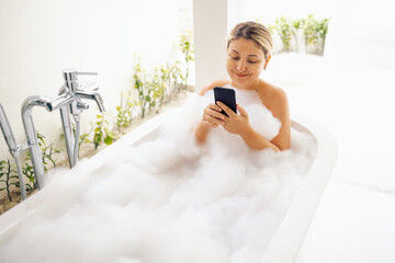 Young woman holds phone and takes bubble bath. Cute blonde is enjoying hot water and chatting on internet