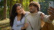 © Yuliia - Glad positive multiethnic couple Hispanic female girlfriend woman Arabian Indian male man boyfriend touch smiling making selfie photo mobile phone talking video call streaming in nature park outdoors