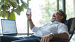 © forenna - proud middle-aged Hispanic man, leaning back in his chair with an elated look, holding his smartphone up in one hand while a laptop displays business graphs on his desk, capturing