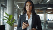© forenna - focused Latin Hispanic female businesswoman holding her smartphone, engaged in online work while standing in a stylish office environment, demonstrating a blend of confidence and t