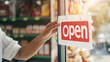 © forenna - Detailed shot of a small business owner adjusting the 'open' sign on a retail store door, reflecting anticipation and readiness to serve customers, with vibrant decor and merchandi