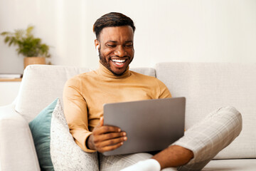 Wall Mural - Cheerful Black Man In Wireless Earphones Using Laptop Computer Watching Movie Sitting On Sofa Indoor. Free Space