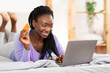 © Prostock-studio - Online Shopping. Afro Lady Using Laptop Showing Credit Card To Camera Purchasing Things On Internet Lying In Bed At Home.
