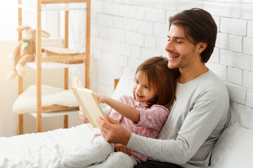 Wall Mural - Little daughter and daddy enjoying weekend together, sitting on bed at home, man reading book to his cute daughter, empty space