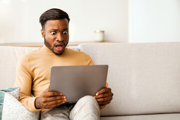 Wall Mural - Shocked Black Guy Looking At Laptop Computer Working Sitting On Couch At Home. Selective Focus