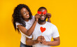© Prostock-studio - Happy Valentine's Day darling. Black pretty woman closing man eyes with red valentine cards on yellow background