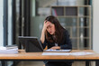 © Wicitr - Overwhelmed and Exhausted: Young Asian businesswoman grapples with work stress, holding her head in her hand at her desk, surrounded by paperwork and digital devices.