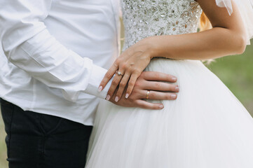 Wall Mural - Groom in white shirt with bracelet and bride in dress with gold ring on finger standing outdoors holding hands. Close-up wedding photo of happy newlyweds.