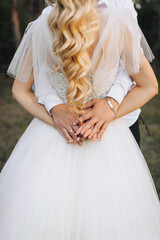 Wall Mural - Groom with bracelet hugging bride with curly hair in white dress from behind holding hands outdoors. Wedding photo of beautiful newlyweds newlyweds.