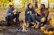 © Prostock-studio - Camping time for mixed race group of friends sitting beside fireplace and frying sausages in autumn forest in sun lights